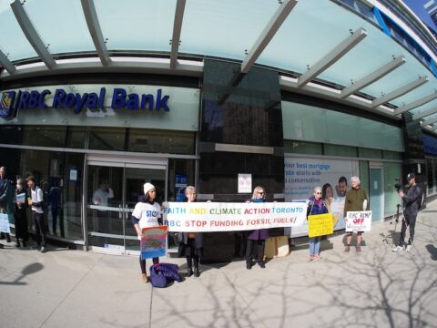 A group of protestors of different genders, ages, ethnicities stand in front of a RBC Bank in Toronto. Three of them are holding a banner that says, "Faith and Climate Action Toronto" on one line and on a second line, it says, "RBC Stop Funding Fossil Fuels!"