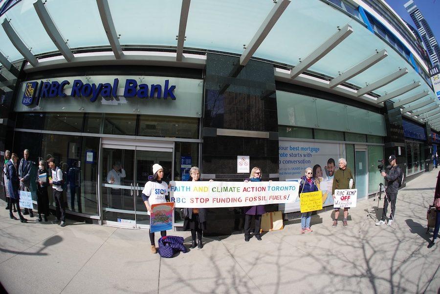 A group of protestors of different genders, ages, ethnicities stand in front of a RBC Bank in Toronto. Three of them are holding a banner that says, "Faith and Climate Action Toronto" on one line and on a second line, it says, "RBC Stop Funding Fossil Fuels!"