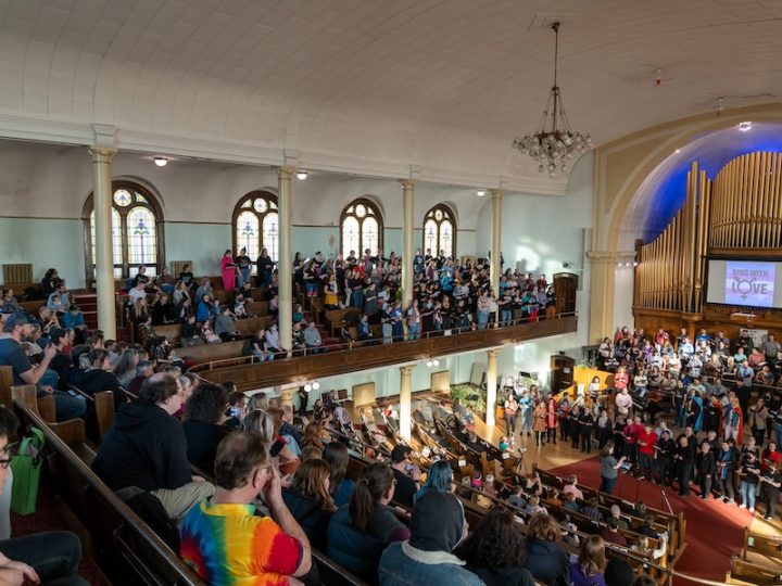 A church is packed with people watching a choir sing. The perspective is from the upper pews. The choir are performing on stage on the lower level in front of an organ. There is a screen in front of the organ. In black text, it says "Sing with Love" on top of a trans flag. The in "O" Love is the trans symbol. The choir is standing and the audience is sitting.