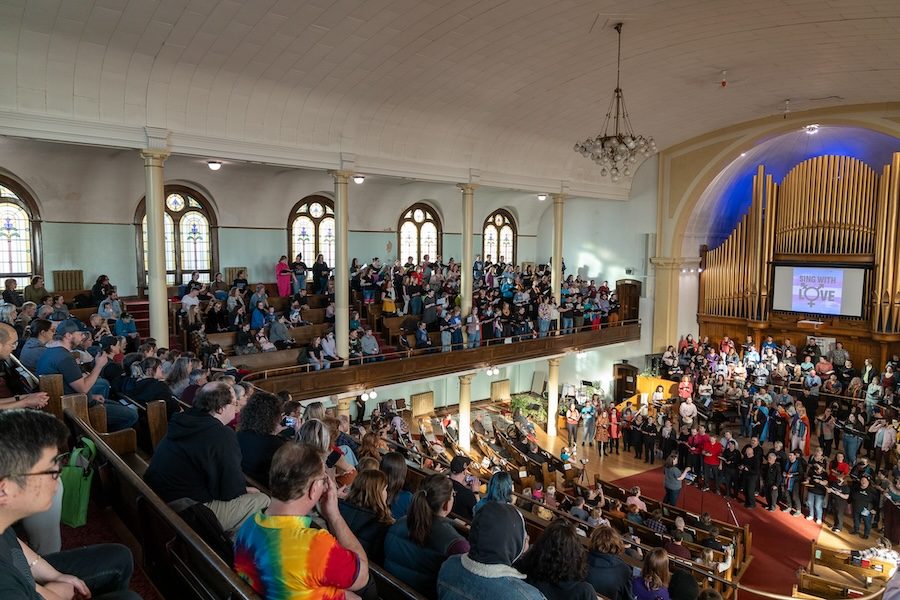 A church is packed with people watching a choir sing. The perspective is from the upper pews. The choir are performing on stage on the lower level in front of an organ. There is a screen in front of the organ. In black text, it says "Sing with Love" on top of a trans flag. The in "O" Love is the trans symbol. The choir is standing and the audience is sitting.