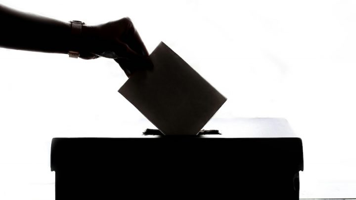 A backlit image of a person putting a vote into a ballot box. The hand holding the ballot and the box are in black and white shadows.