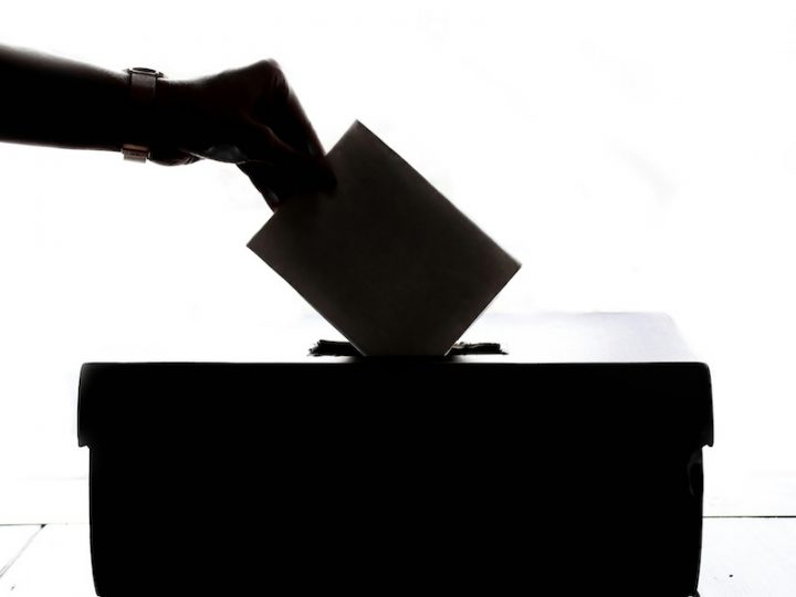 A backlit image of a person putting a vote into a ballot box. The hand holding the ballot and the box are in black and white shadows.