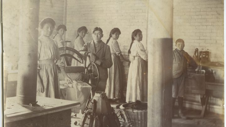 A sepia-toned colour, grainy, old photo from 1909 featuring a group of Indigenous pupils at Mount Elgin Industrial Institute, a United Church-run residential school in Ontario. The students are doing laundry using old washing wheels. The girls have long braids, long-sleeve shirts and skirts and aprons on. The boys have short hair and blazers and trousers on.