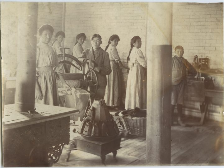 A sepia-toned colour, grainy, old photo from 1909 featuring a group of Indigenous pupils at Mount Elgin Industrial Institute, a United Church-run residential school in Ontario. The students are doing laundry using old washing wheels. The girls have long braids, long-sleeve shirts and skirts and aprons on. The boys have short hair and blazers and trousers on.