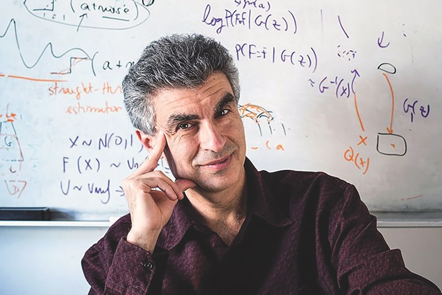 A middle-aged man with white skin, salt and pepper hair poses. He is in front of a white board with equations. His face is resting on his right hand and he is wearing a purple button-up shirt. 