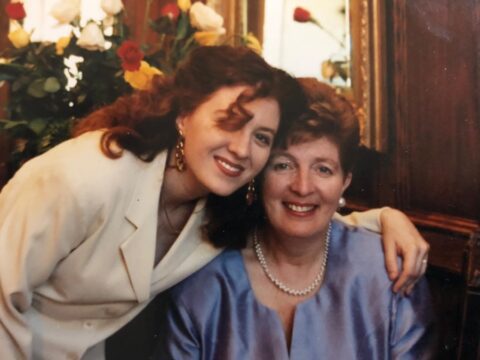 The author, Allyson McOuat poses with her mother. Allyson is in a cream-coloured suit, with reddish brown, shoulder-length curly hair and red lipstick. She is smiling and has her arm around her mother. Her mother is wearing a light blue dress, white pearls, short reddish-brown hair and is also smiling.
