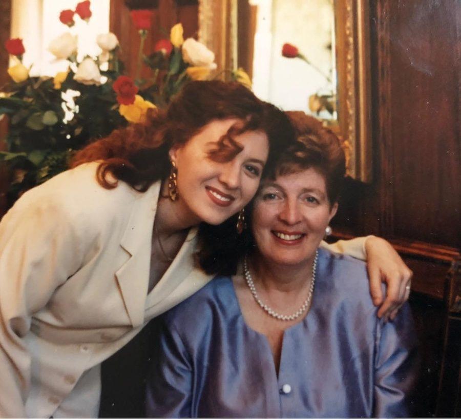 The author, Allyson McOuat poses with her mother. Allyson is in a cream-coloured suit, with reddish brown, shoulder-length curly hair and red lipstick. She is smiling and has her arm around her mother. Her mother is wearing a light blue dress, white pearls, short reddish-brown hair and is also smiling.