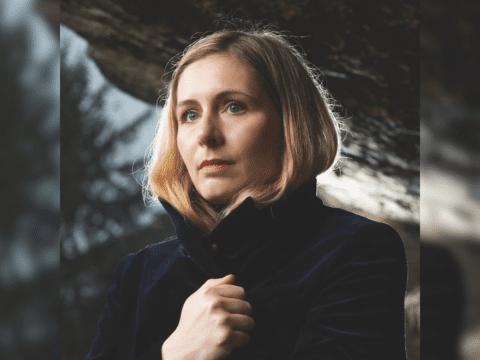 A blond, white woman with blue eyes poses. She has short blond hair, her hand is holding her black coat closed.