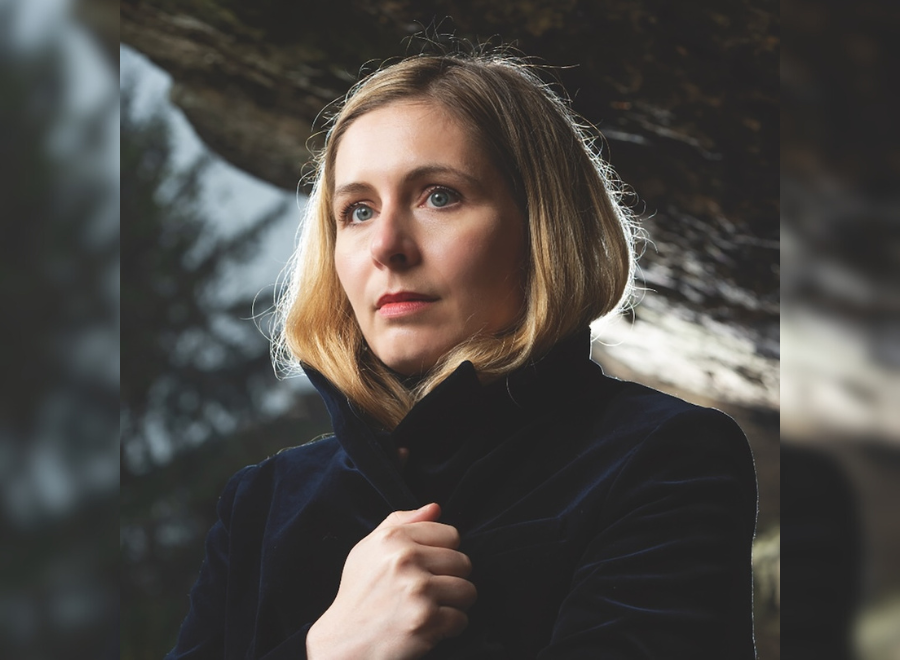 A blond, white woman with blue eyes poses. She has short blond hair, her hand is holding her black coat closed.