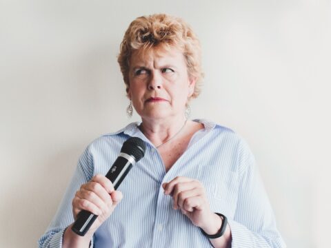 A middle-aged white woman with short curly blonde hair is holding a microphone and looking dismayed. She is wearing a blue and white striped shirt with a silver necklace and silver dangling earrings. She has on red lipstick.