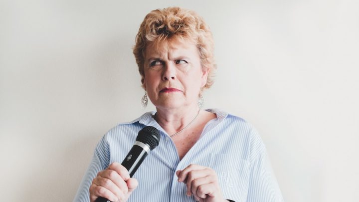 A middle-aged white woman with short curly blonde hair is holding a microphone and looking dismayed. She is wearing a blue and white striped shirt with a silver necklace and silver dangling earrings. She has on red lipstick.