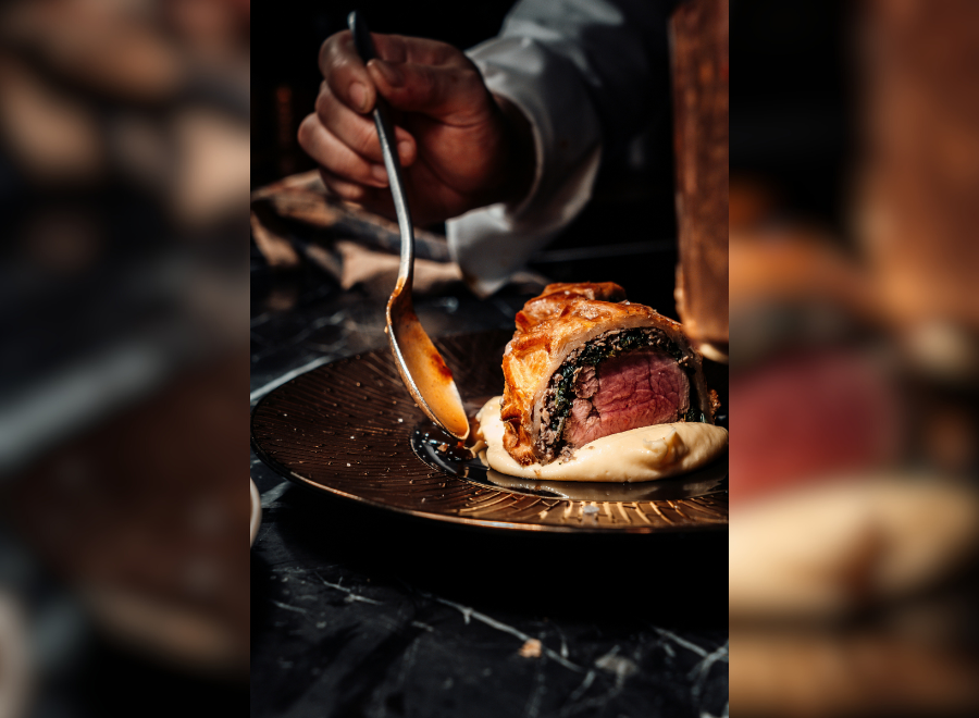 A close up image of a piece of steak being plated. A Black hand is holding a spoon that is dipping into the sauce next to the steak.