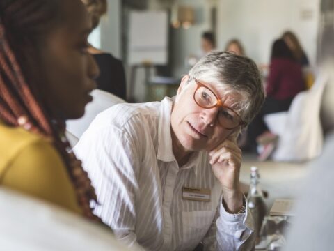A middle-aged white woman with short hair is wearing brown-framed glasses and talking to someone off frame. She is wearing a white striped button-up shirt. Her chin is resting on her left hand. She is wearing a nametag that says Ellen Woodsworth.