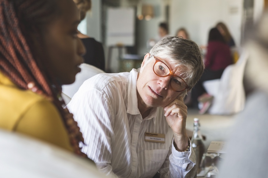 A middle-aged white woman with short hair is wearing brown-framed glasses and talking to someone off frame. She is wearing a white striped button-up shirt. Her chin is resting on her left hand. She is wearing a nametag that says Ellen Woodsworth.