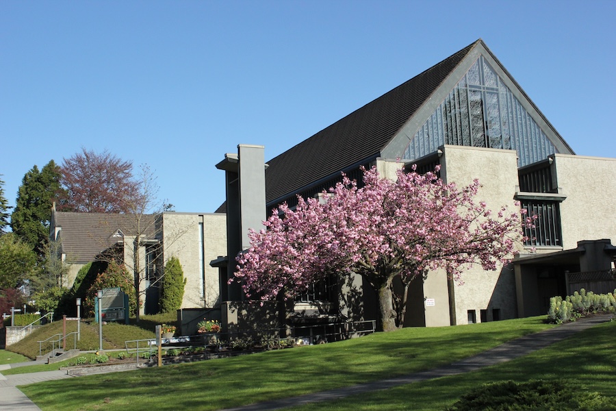 Image of a church in British Columbia. In front of the church is a tree full of pink blossoms and a green lawn and picnic table.