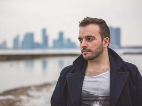 A white-skinned man stands in front of a city line and a body of water. He has brown hair and is wearing a navy jacket with a white shirt underneath. He has a scruffy beard.