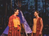 Two Indigenous women are standing in front of a purple tipi. The women are dressed in red and yellow dresses. One of the dresses has full sleeves and the other is sleeveless. One woman has dark brown hair tied into a braid and the other woman has light brown hair in a bun. They are talking and smiling.