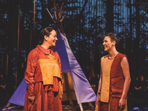 Two Indigenous women are standing in front of a purple tipi. The women are dressed in red and yellow dresses. One of the dresses has full sleeves and the other is sleeveless. One woman has dark brown hair tied into a braid and the other woman has light brown hair in a bun. They are talking and smiling.
