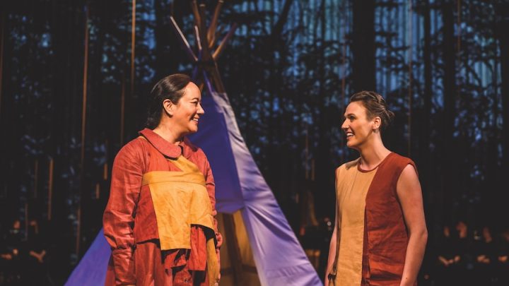 Two Indigenous women are standing in front of a purple tipi. The women are dressed in red and yellow dresses. One of the dresses has full sleeves and the other is sleeveless. One woman has dark brown hair tied into a braid and the other woman has light brown hair in a bun. They are talking and smiling.