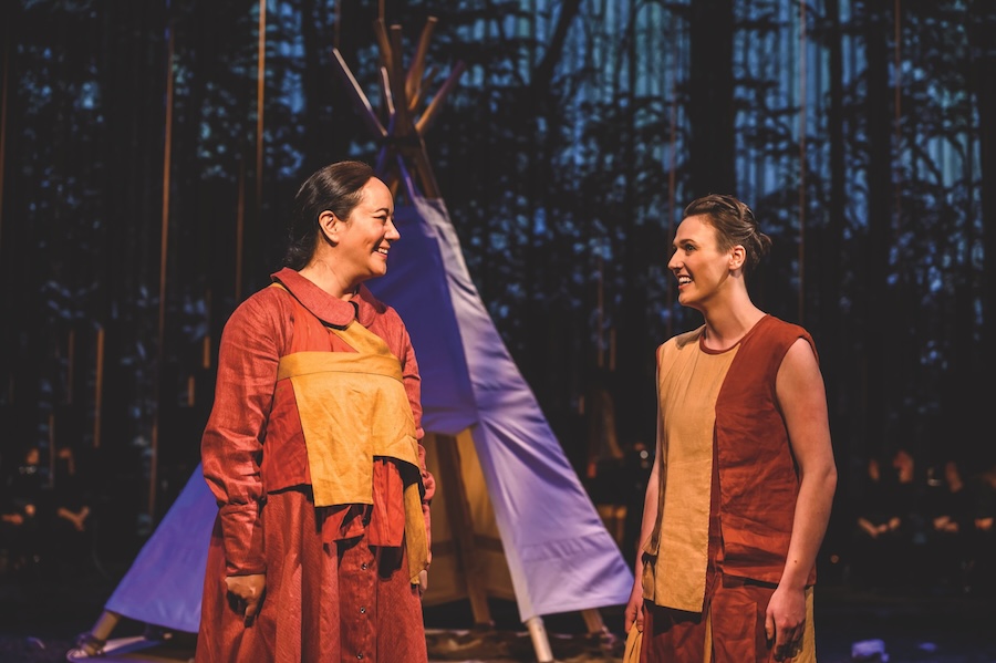 Two Indigenous women are standing in front of a purple tipi. The women are dressed in red and yellow dresses. One of the dresses has full sleeves and the other is sleeveless. One woman has dark brown hair tied into a braid and the other woman has light brown hair in a bun. They are talking and smiling.
