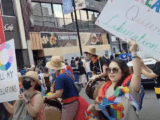 Image depicts a crowd of people. At the front, a woman holds a sign reading "Queer Liberation" and carries rainbow objects. To her left is a person wearing a rainbow scarf and holding a drum. To their left is a masked woman holding up another sign.