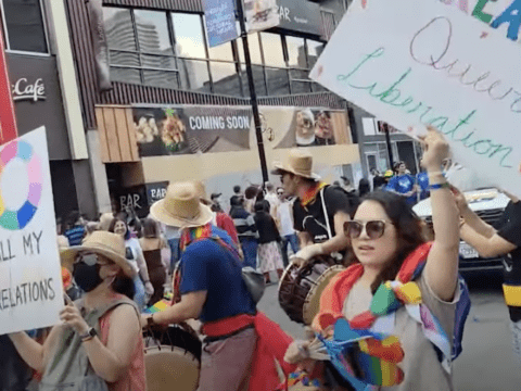 Image depicts a crowd of people. At the front, a woman holds a sign reading "Queer Liberation" and carries rainbow objects. To her left is a person wearing a rainbow scarf and holding a drum. To their left is a masked woman holding up another sign.