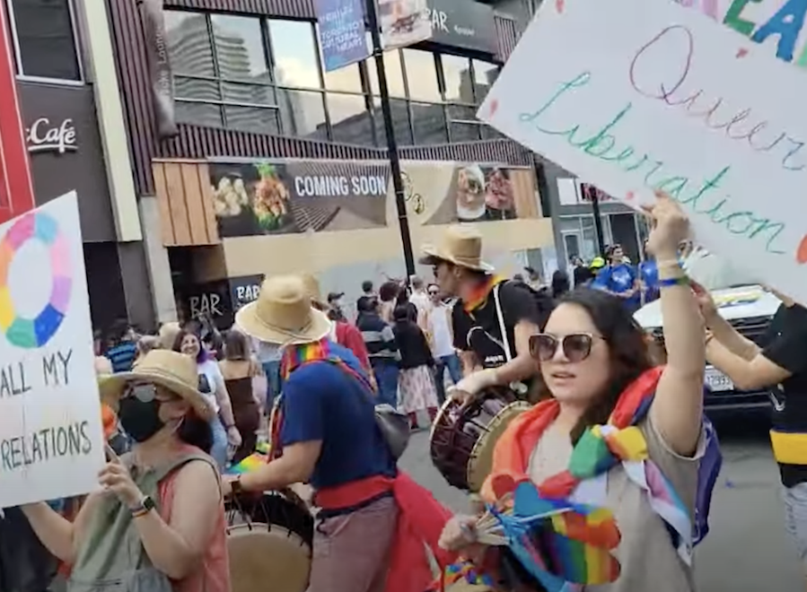 Image depicts a crowd of people. At the front, a woman holds a sign reading "Queer Liberation" and carries rainbow objects. To her left is a person wearing a rainbow scarf and holding a drum. To their left is a masked woman holding up another sign.