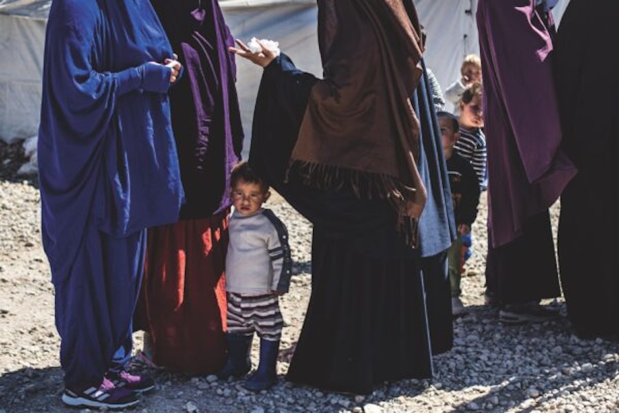 Small children, including one wearing rubber boots and striped pants, stand amid adults on rocky ground.