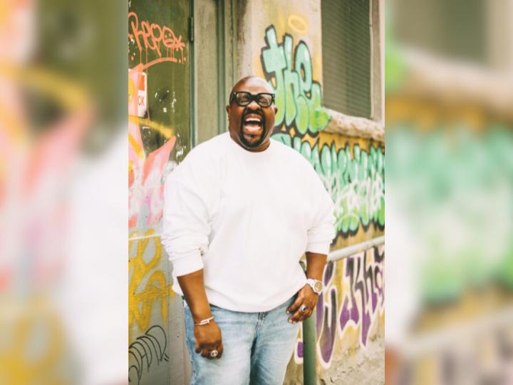 A bald Black man with black-framed glasses is standing in front of graffiti and laughing with his eyes closed and mouth open. He has a goatee and earrings in each ear. His left hand is in the pocket of his blue jeans. he is wearing a white sweatshirt. On his right hand is a white watch with a ring on his middle finger. He is also wearing a ring on the left middle finger and a silver bracelet.
