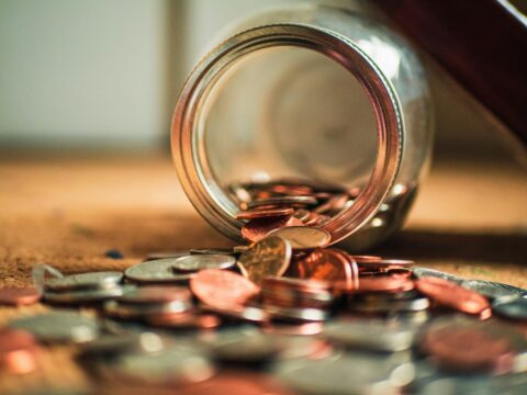 An open jar of coins is on a surface and half of the coins have spilled out.