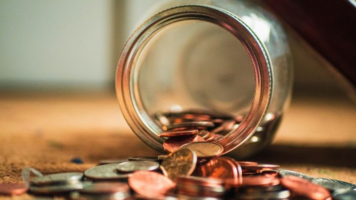 An open jar of coins is on a surface and half of the coins have spilled out.