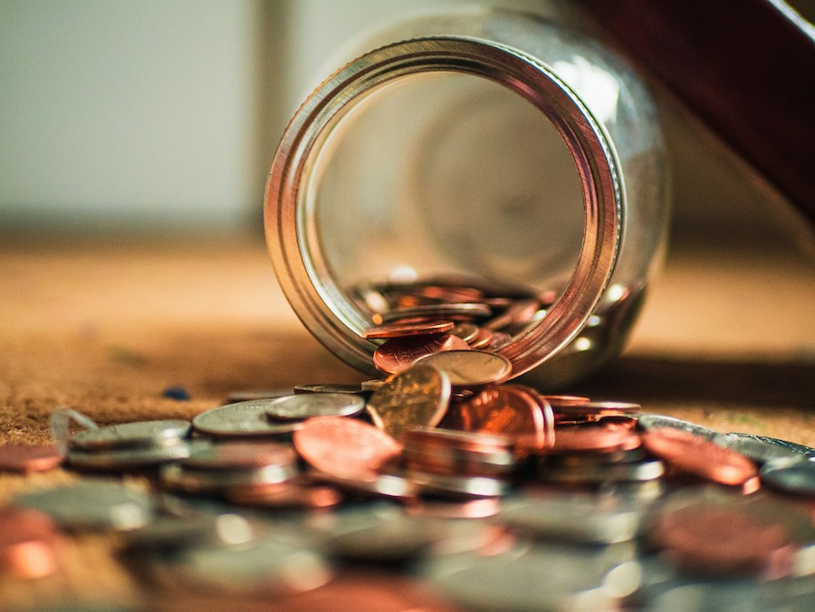 An open jar of coins is on a surface and half of the coins have spilled out.