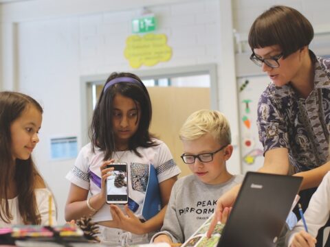 A Finnish teacher teaches a group of students. The teacher is wearing black-framed glasses and has a bowl cut with short brown hair and bangs. She is wearing a paisley button-up shirt and pointing at a book. Next to her is a blond boy with black-framed glasses, wearing a grey sweatshirt. Next to him is a brown-skinned girl with shoulder-length brown hair and wearing a purple headband. She has on a white t-shirt and is holding up a mobile phone with an image of an acorn. Beside her is a brown-skinned girl with long brown hair. She is wearing a white shirt and looking at the book.