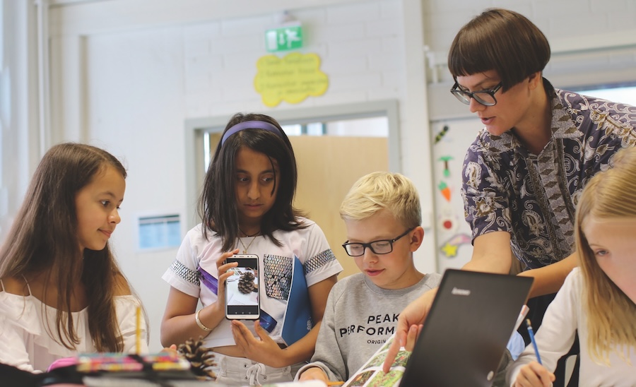 A Finnish teacher teaches a group of students. The teacher is wearing black-framed glasses and has a bowl cut with short brown hair and bangs. She is wearing a paisley button-up shirt and pointing at a book. Next to her is a blond boy with black-framed glasses, wearing a grey sweatshirt. Next to him is a brown-skinned girl with shoulder-length brown hair and wearing a purple headband. She has on a white t-shirt and is holding up a mobile phone with an image of an acorn. Beside her is a brown-skinned girl with long brown hair. She is wearing a white shirt and looking at the book.