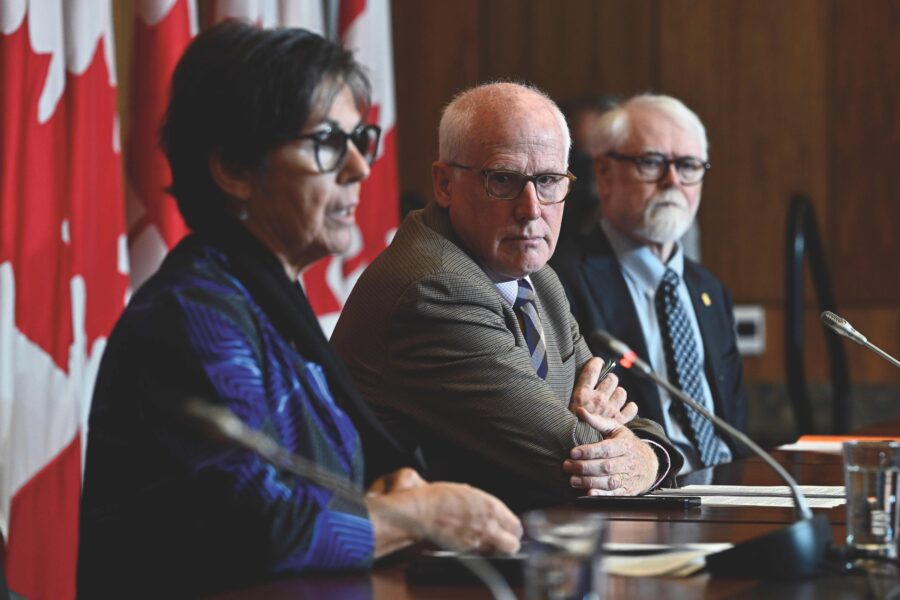 Three people are speaking in front of microphones and they are seated. From left to right: A woman with glasses, short hair and a blue shirt. Next to her is a bald, white man with glasses in a grey suit. Next to him is an older white man with white hair, white facial hair, glasses and a black suit and tie on