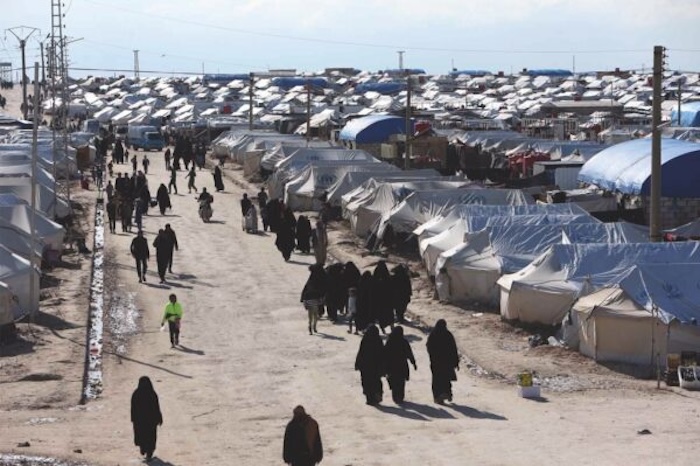 Women in head coverings walk down a dirt path next to a sea of tents.
