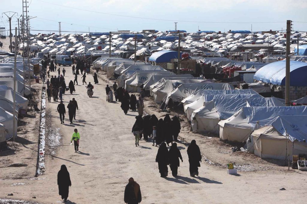 Women in head coverings walk down a dirt path next to a sea of tents. 