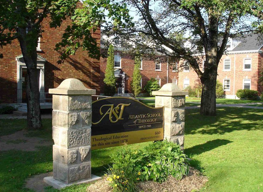 A sign reading "Atlantic School of Theology" sits on the grass, in front of brick buildings.