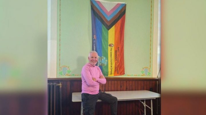 A man in a pink sweater, Kevin Moore, leans on a folding table in front of a 2SLGBTQ+ Pride Flag.