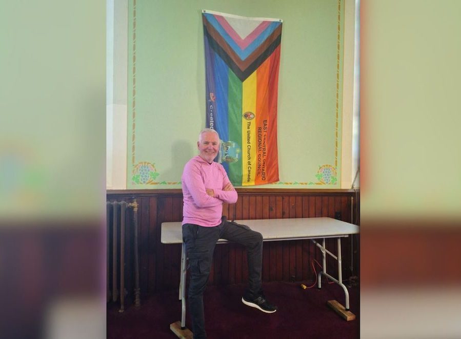 A man in a pink sweater, Kevin Moore, leans on a folding table in front of a 2SLGBTQ+ Pride Flag.