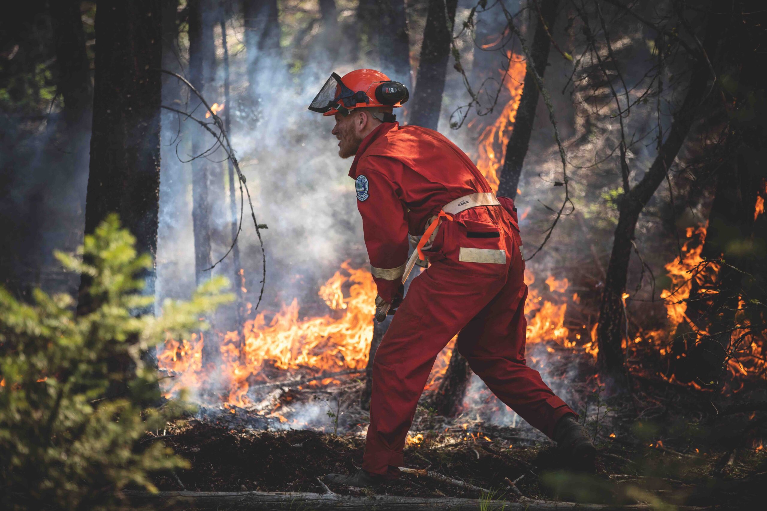 A firefighter wearing a helmet and dressed in an orange jumpsuit faces a blazing forest fire.