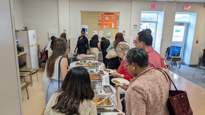 A group of people are lined up around a table of food, helping themselves. The perspective is from behind so you cannot see people's faces but it is a diverse group of women with different hair colours and styles wearing a variety of shirts and outfits.