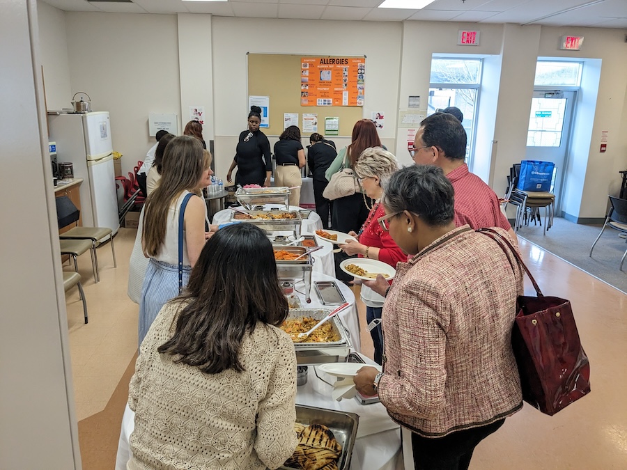 A group of people are lined up around a table of food, helping themselves. The perspective is from behind so you cannot see people's faces but it is a diverse group of women with different hair colours and styles wearing a variety of shirts and outfits.