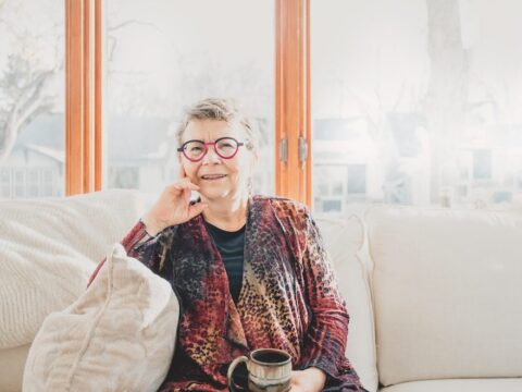 A woman, Janis Hughes, sits on a couch in front of a window, holding a mug.