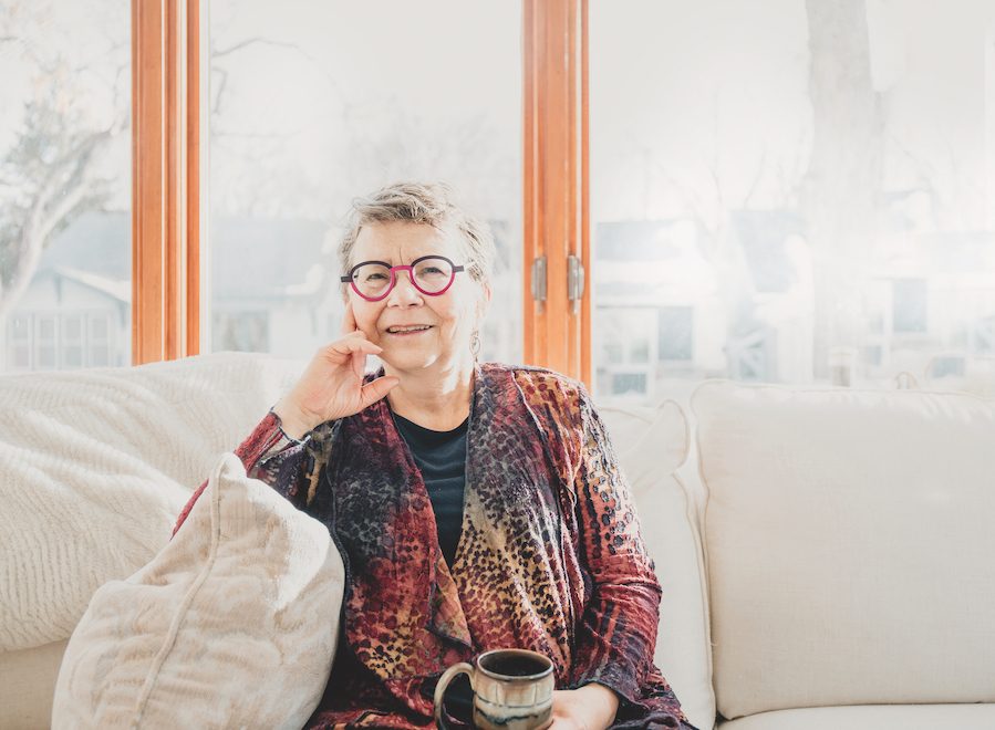 A woman, Janis Hughes, sits on a couch in front of a window, holding a mug.