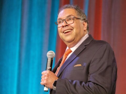 A middle-aged brown-skinned man with short curly brown and grey hair holds a microphone in his left hand. He is wearing black-framed glasses, a white button up shirt, red tie and a navy suit.