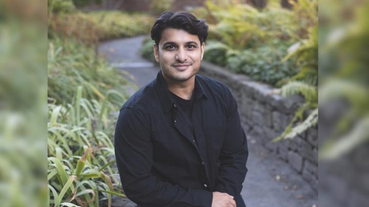 A brown-skinned man with short dark hair, in a long-sleeved black shirt and black pants sits in front of a garden. There are green grasses behind him.