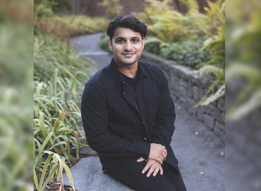 A brown-skinned man with short dark hair, in a long-sleeved black shirt and black pants sits in front of a garden. There are green grasses behind him.