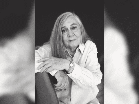 A middle-aged white woman with grey hair poses on a chair. The photo is in black and white. She is wearing a light-coloured button up and a watch on her left hand. Her left hand rests on her right arm.