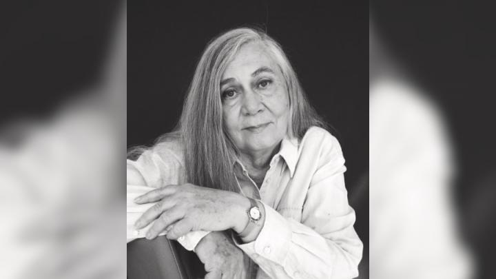 A middle-aged white woman with grey hair poses on a chair. The photo is in black and white. She is wearing a light-coloured button up and a watch on her left hand. Her left hand rests on her right arm.